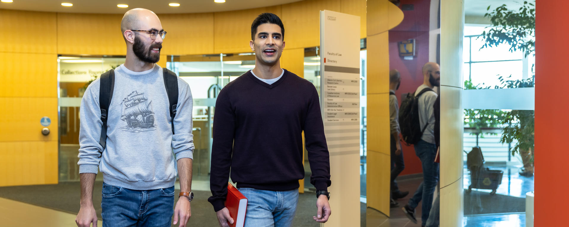 Two students walk through a hallway. One is wearing a grey sweatshirt and is carrying a backpack. The other is wearing a black sweater and is carrying a book.